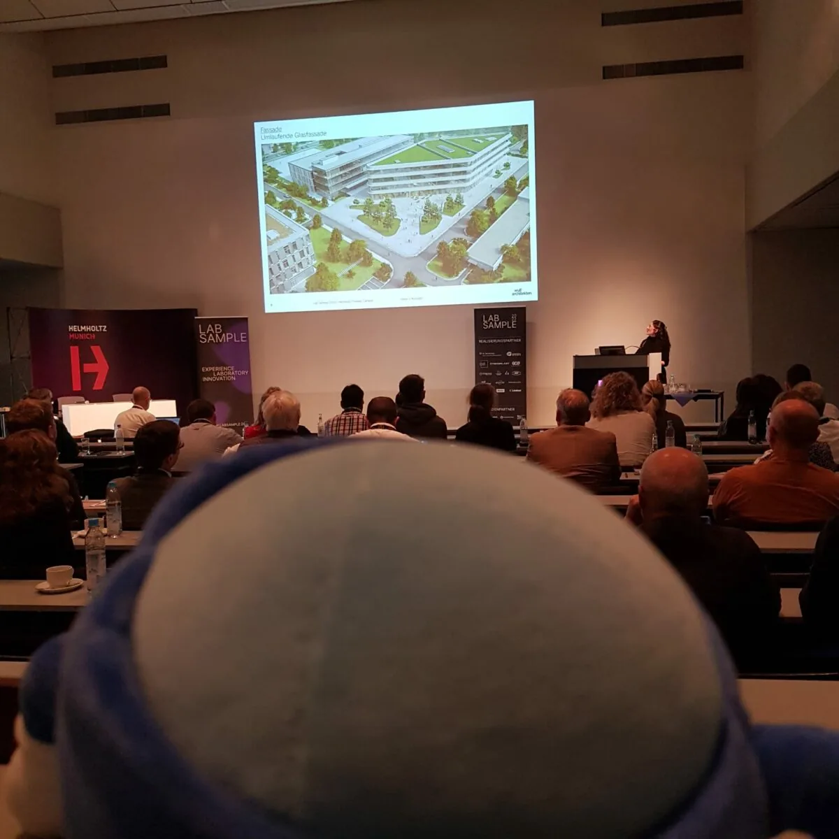The image shows a conference or seminar setting with an audience seated and facing a presenter. The