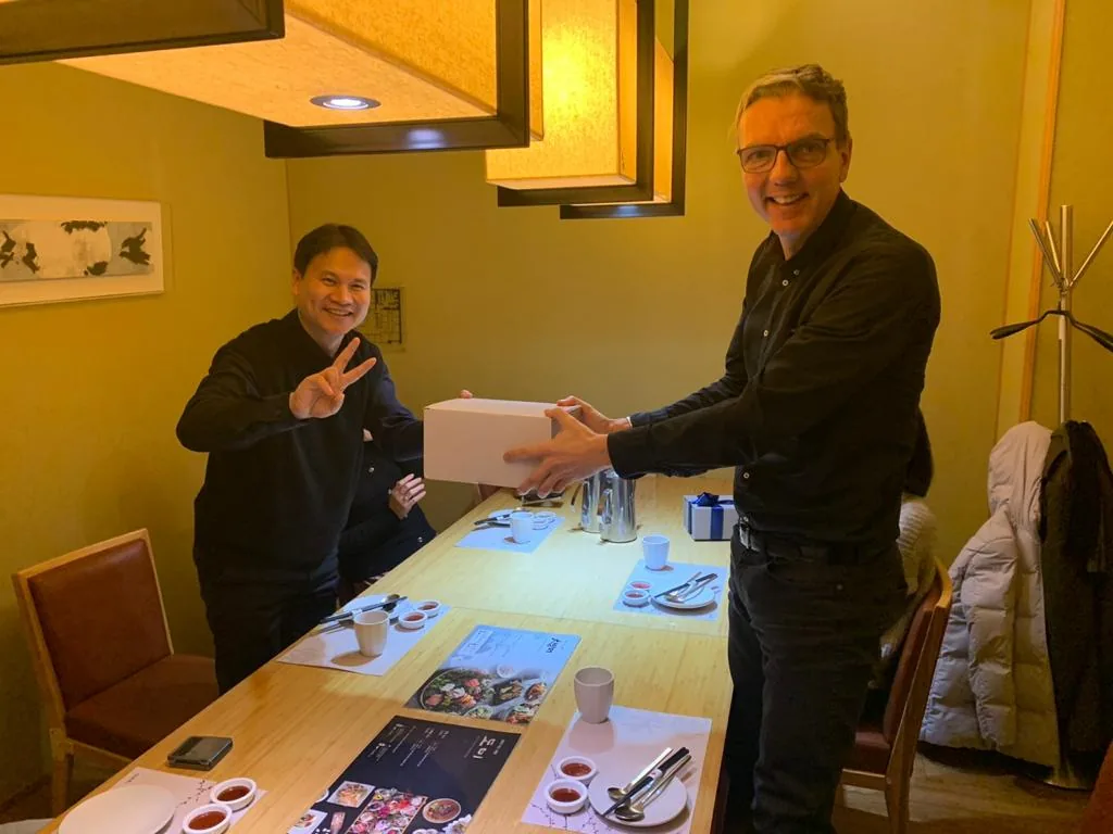 Two men are smiling at a dining table, one holding a white box while the other makes a peace sign